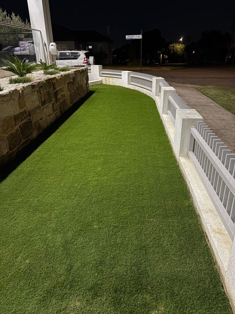 A curved front yard lawn with artificial turf, stone retaining wall, and white fence completed during landscaping at night.